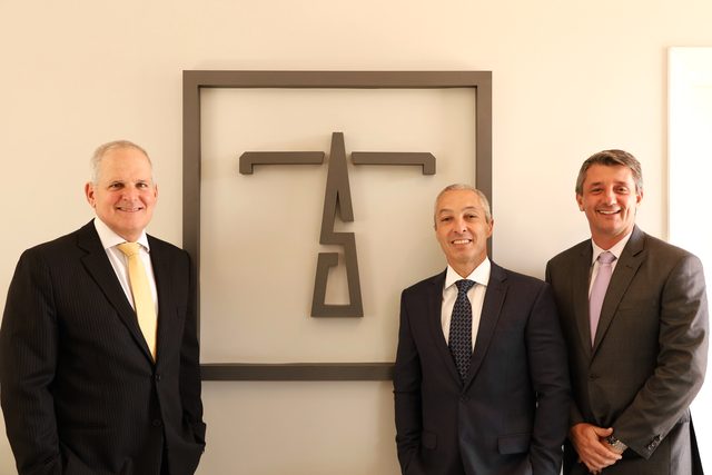 Os sócios Gustavo Moura Tavares, Fernando Cesar Barbosa Siqueira e Matheus de Toledo diante do monograma do escritório, na sede